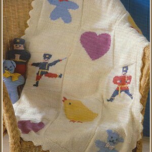 May include: A cream-colored knitted blanket featuring a grid of square panels with intarsia designs including blue teddy bears with bows, a purple heart, a yellow chick, and two figures dressed in blue and red military-style uniforms.