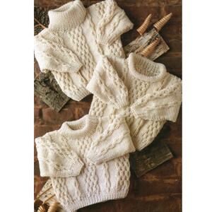 May include: Three cream-coloured knitted jumpers with a cable knit pattern. The jumpers are folded and arranged on a wooden surface.