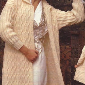 May include: Cream-colored, long cable-knit cardigan with a shawl collar. The cardigan is worn over a white collared shirt and a layered necklace. The sleeves are long, and the cardigan reaches the ankles. The knitwear has a textured pattern.