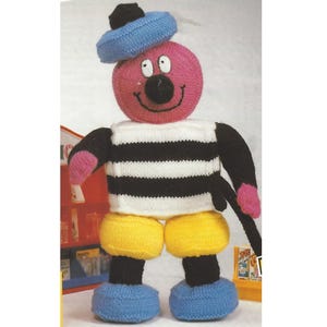 May include: A knitted toy with a pink and white head, black and white striped torso, yellow trousers, and blue shoes. The toy is wearing a blue hat.