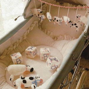 May include: A white baby carriage with a crocheted mobile hanging above the mattress. The mobile features five crocheted white and black spotted cows. The carriage also has four crocheted blocks with letters on them. There are two crocheted white and black spotted cows on the mattress.