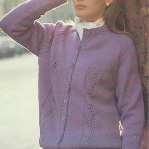 Könnte beinhalten: Ein fliederfarbener Cardigan mit rundem Ausschnitt und Knopfleiste. Der Strickpullover hat ein zartes Blättermuster auf der Vorderseite und den Ärmeln. Das Model trägt eine weiße Bluse darunter und einen grauen Rock.