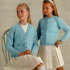 Peut inclure: Deux filles portant des cardigans bleu clair avec des boutons. La fille de gauche porte une jupe blanche et la fille de droite porte une jupe plissée blanche.