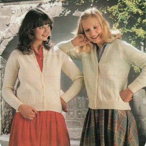 Puede incluir: Dos mujeres con cárdigans color crema abotonados. Una mujer lleva un vestido rojo, la otra una falda a cuadros. Los cárdigans tienen mangas largas y un dobladillo acanalado. El fondo presenta un muro de piedra y vegetación.