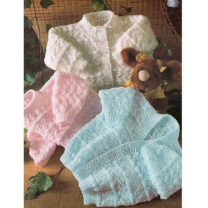 May include: Three knitted baby sweaters in pink, light blue, and white with a textured pattern. The sweaters are laid out on a wooden surface with green leaves.