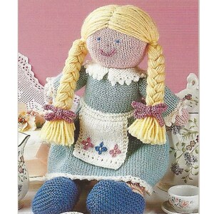 May include: A knitted doll with blonde braids, wearing a blue dress with a white apron and pink bows. The doll is sitting on a white surface with a pink background.