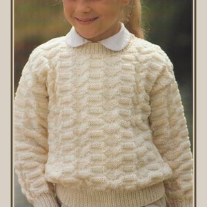 May include: A cream-coloured knitted jumper with a textured pattern. The jumper has a ribbed hem and cuffs, and a white collared shirt is visible underneath. The jumper is worn by a person.
