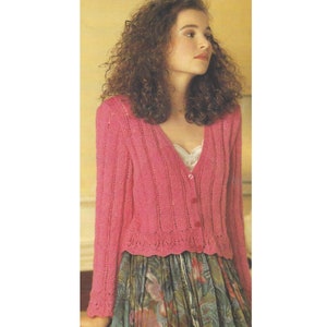 Może przedstawiać: Różowy kardigan z dzianiny z dekoltem w kształcie litery V i zapięciem na guziki. Kardigan ma koronkową lamówkę na dole i na rękawach. Modelka ma na sobie biały top.