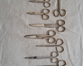May include: A collection of 11 surgical scissors of varying sizes and styles, laid out on a white fabric surface.