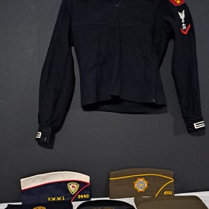 May include: A black wool jacket with white trim and various patches, including a red eagle, a white dove, and a red poppy.  There are also three hats: a blue and white hat with the text "V.W.W.1 3440", a green hat with the text "4701", and a black hat with the text "U.S. NAVY".