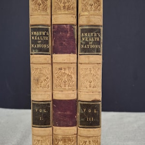 May include: Three antique books bound in tan leather with gold accents and burgundy spines. The books are titled "Smith's Wealth of Nations" and are labelled "Vol. I", "Vol. II", and "Vol. III".