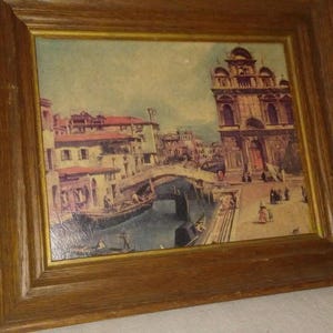 以下が含まれることがあります： イタリアのヴェネツィアの運河のシーンを描いた額装された絵画。絵画には橋、ゴンドラ、大きな教会を背景にした建物が描かれている。