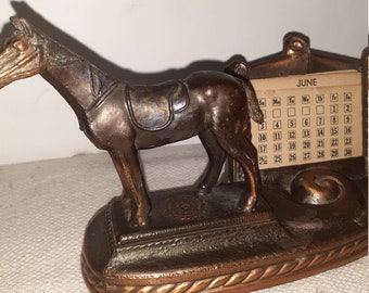 May include: A vintage bronze desk calendar featuring a horse figurine. The calendar is for the month of June and has a small, round, decorative base.