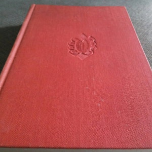 Puede incluir: Un libro de tapa dura rojo con un diseño en relieve en la portada. El diseño es una imagen estilizada de una flor.