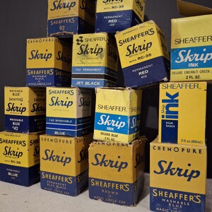 May include: A collection of vintage Sheaffer's Skrip ink boxes in various colors, including red, blue, and black. The boxes are yellow and blue, with the Skrip logo prominently displayed. Some boxes indicate the ink type, such as "Washable Blue" and "Permanent Red".
