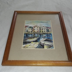 Puede incluir: Una pintura al agua enmarcada que representa un pequeño pueblo con edificios coloridos a lo largo de una costa. Dos barcos están atracados en el agua, y la escena está pintada en un estilo suelto e impresionista.