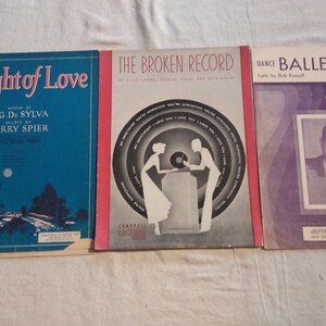 May include: Three vintage sheet music covers. The first cover is blue with a night scene and the title "A Night of Love" by B.G. De Sylva and Larry Spier. The second cover is red with a black and white illustration of a couple listening to a record player and the title "The Broken Record". The third cover is purple with a black and white photo of a man and the title "Dance Ballerina Dance" by Bob Russell and Carl Sigman.