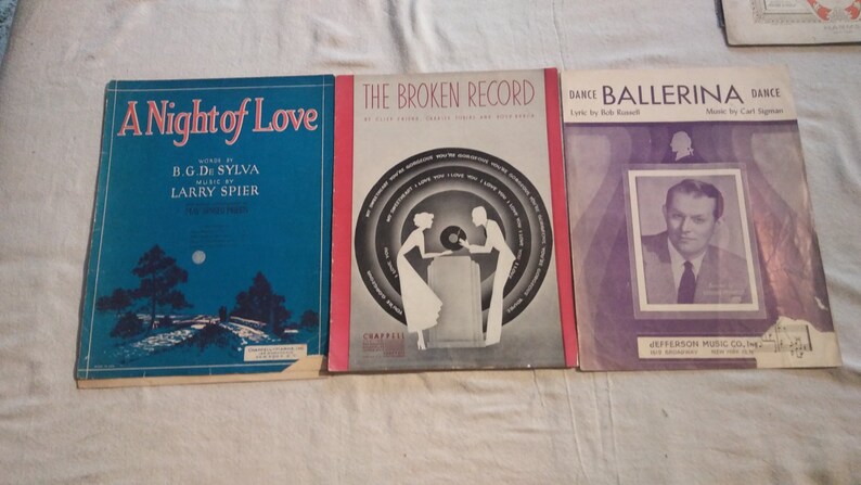 May include: Three vintage sheet music covers. The first cover is blue with a night scene and the title "A Night of Love" by B.G. De Sylva and Larry Spier. The second cover is red with a black and white illustration of a couple listening to a record player and the title "The Broken Record". The third cover is purple with a black and white photo of a man and the title "Dance Ballerina Dance" by Bob Russell and Carl Sigman.