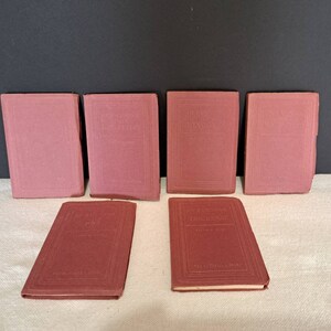 May include: Seven small, burgundy-coloured books with gold lettering on the spines. The books are titled "The Writings of Thomas Carlyle" and "The Writings of Thackeray".