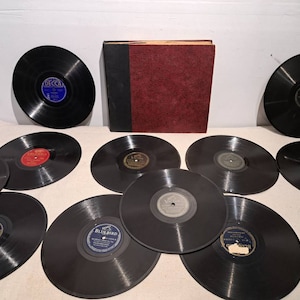 Peut inclure: Une collection de disques vinyles vintage, dont plusieurs avec des étiquettes bleues et rouges, et un étui d'album de couleur bordeaux. Les disques sont noirs et disposés sur une surface blanche, mettant en valeur le design classique des étiquettes.