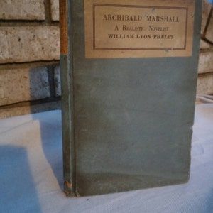 Peut inclure: Un livre vert avec une étiquette jaune sur laquelle est écrit "Archibald Marshall A Realistic Novelist William Lyon Phelps".