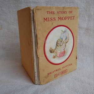 May include: A vintage book titled "The Story of Miss Moppet" by Beatrix Potter. The cover features a colorful illustration of a cat holding a bunch of blue flowers. The book is published by F. Warne & Co.