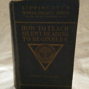 Puede incluir: Un libro azul titulado "How to Teach Silent Reading to Beginners" de la serie Lippincott's School Project Series, editado por W.F. Russell, Ph.D. El libro presenta un triángulo dorado con un escudo y el texto "DROIT ET AVANT" en su interior. La autora es Emma Watkins.