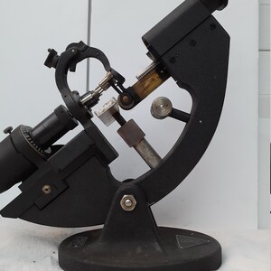 Puede incluir: Un instrumento oftalmológico de metal negro con una base circular y un brazo largo y curvo. El instrumento tiene una variedad de engranajes y palancas, y una pequeña lente tipo telescopio en el extremo del brazo.