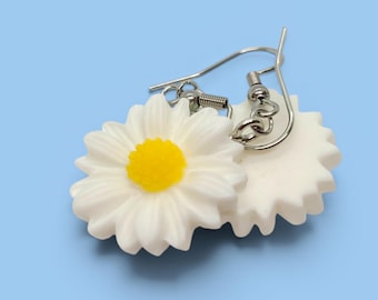 May include: A pair of daisy earrings with white petals and a yellow center. The earrings have a silver-colored hook and are set against a light blue background. The earrings are a floral-themed accessory.