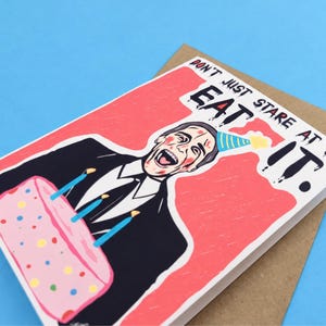 May include: A greeting card featuring a cartoon illustration of a person in a suit and party hat holding a pink cake with lit candles. The text reads, "DON'T JUST STARE AT IT. EAT IT." The card is placed on top of a brown envelope.