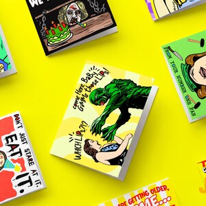 May include: A collection of rectangular greeting cards featuring colorful pop-art style illustrations. Designs include a green creature reaching for a person, a person with a party hat, and various text captions like EAT IT and GIMME those Lips.