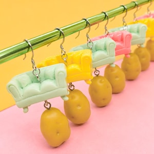 May include: A collection of novelty earrings featuring miniature colorful sofas and potato charms. The earrings are in various colors, including mint green, yellow, and pink, hanging from a green metal bar against a pink and yellow background.