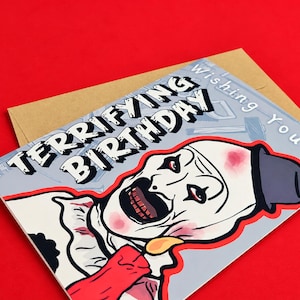 May include: A rectangular greeting card with a brown envelope featuring a graphic of a clown with white face paint, a black hat, and red accents. The text reads "Wishing You A TERRIFYING BIRTHDAY" in bold black and white lettering.