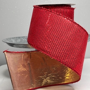 May include: A roll of red sequined ribbon with a gold metallic underside. The ribbon is wide and appears to be unwinding, revealing the shiny gold material. The ribbon is likely used for crafting, gift wrapping, or holiday decorations.