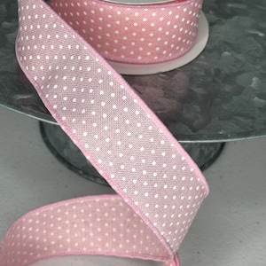 May include: A spool of pink ribbon with white polka dots. The ribbon is made of a woven fabric and is partially unrolled, revealing the pattern. The ribbon is on a metal stand.