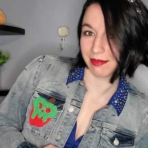 Può includere: Giacca di jeans color azzurro chiaro con colletto blu decorato con borchie argentate. La giacca presenta una toppa a forma di mela all'uncinetto rossa e verde sul petto. Sotto è indossata una camicia blu.
