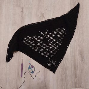 May include: A black crochet shawl with a butterfly design. The shawl is triangular and features a detailed openwork pattern. A crochet hook, scissors, and black yarn are next to the shawl.