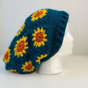 May include: A teal crocheted hat adorned with yellow and orange sunflowers. The hat is displayed on a white mannequin head, showcasing the handmade details and floral design. The hat is a unique accessory.