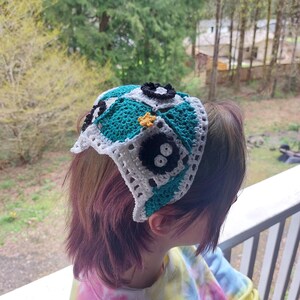 May include: A handmade crochet headband with a patchwork design in teal, white, and black. The headband has a square pattern with flower and star embellishments. The accessory is worn on the head.