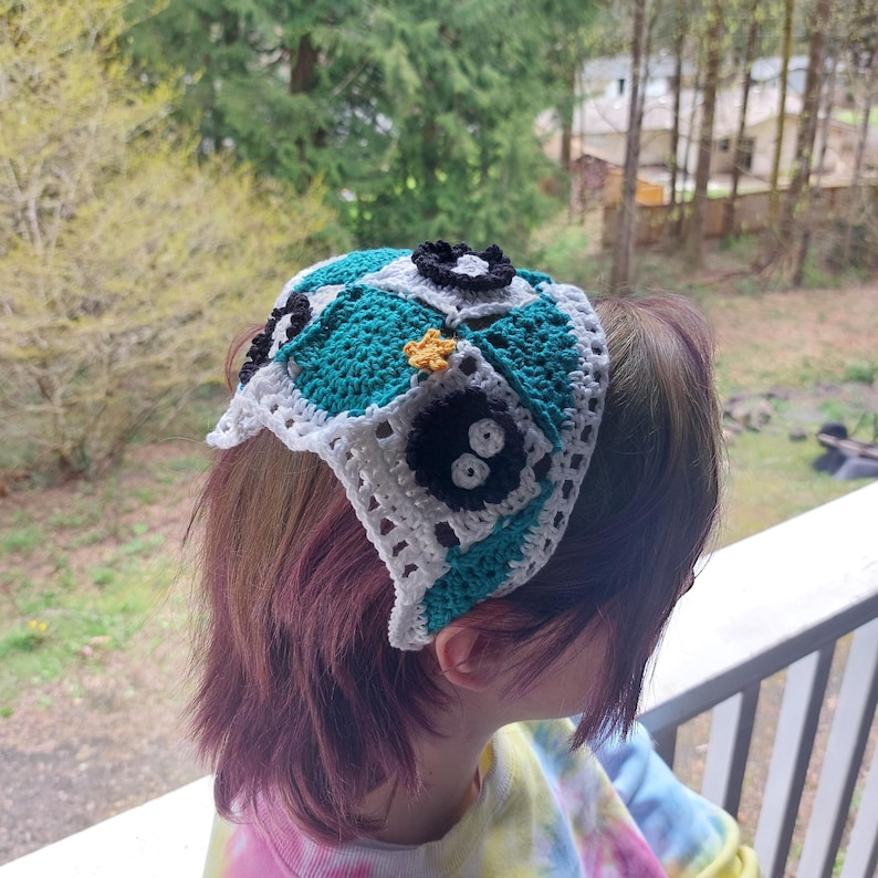 May include: A handmade crochet headband with a patchwork design in teal, white, and black. The headband has a square pattern with flower and star embellishments. The accessory is worn on the head.