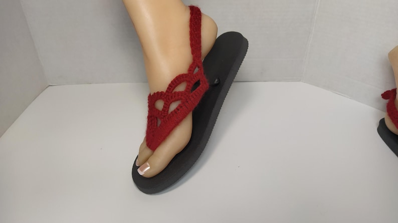 May include: Red crocheted sandals with a black sole. The sandals have a delicate, lacy design and a strap that goes around the ankle.