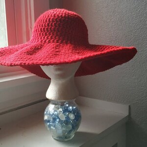 May include: A red crocheted sun hat with a wide brim, sitting on a white mannequin head. The mannequin is on a clear glass jar filled with blue and white glass beads.