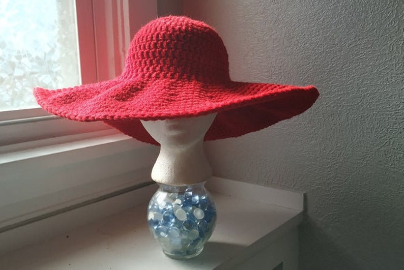 May include: A red crocheted sun hat with a wide brim, sitting on a white mannequin head. The mannequin is on a clear glass jar filled with blue and white glass beads.