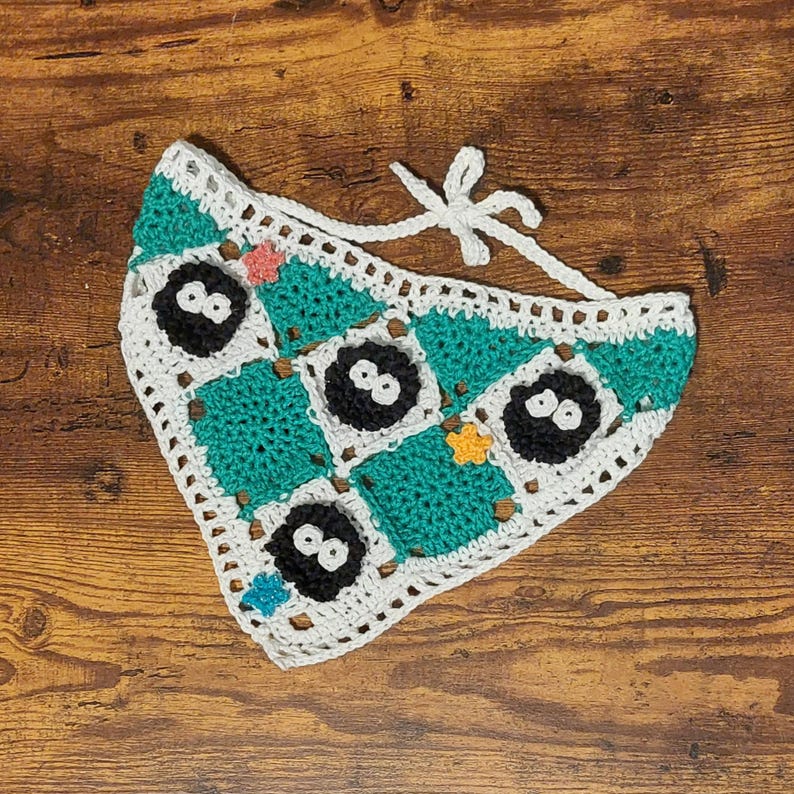 May include: Handmade crochet pet bandana with a checkerboard pattern of teal and white squares. Each teal square has a black soot sprite with white eyes. The bandana has a white border and a white tie.