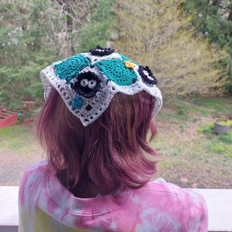May include: A handmade crochet head covering with a square design. The head covering features a white border, teal and black squares, and decorative black and yellow floral accents. The head covering is worn over pink hair.