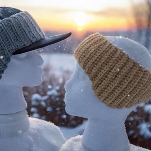 Pode incluir: Duas cabeças de manequim, uma usando um boné de beisebol cinza com protetores de ouvido de malha e a outra uma faixa de cabeça de malha bege. A cena é ambientada em uma paisagem de inverno nevada com um pôr do sol ao fundo.