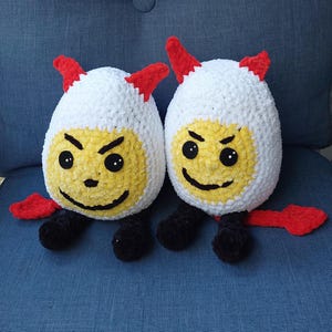Op de afbeelding: Twee handgemaakte, pluche eivormige speelgoedjes. Elk heeft een witte bovenkant met rode hoorns, een gele gezicht met een zwarte boze uitdrukking, zwarte poten en een rode staart. De speeltjes staan op een blauw oppervlak.
