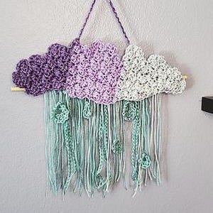 May include: A handmade crochet cloud wall hanging in shades of purple and gray, with teal fringe and decorative accents. The cloud is suspended from a wooden dowel and hangs from a gold hook on a gray wall. A unique piece of home decor.