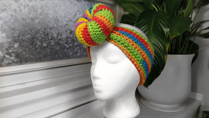 Peut inclure: Un bandeau en crochet color&eacute; avec un gros n&oelig;ud arc-en-ciel sur le dessus. Le bandeau est fait de plusieurs couleurs vives, dont le rouge, l'orange, le jaune, le vert, le bleu et le violet.