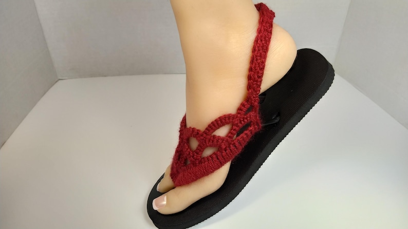 May include: Red crocheted sandals with a black sole. The sandals have a strap that goes around the ankle.